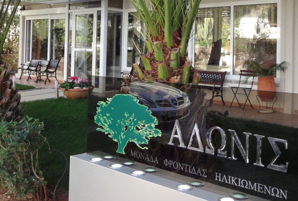Η είσοδος της μονάδας ΑΔΩΝΙΣ με γυάλινη πινακίδα, φοίνικα και καθιστικό στην αυλή